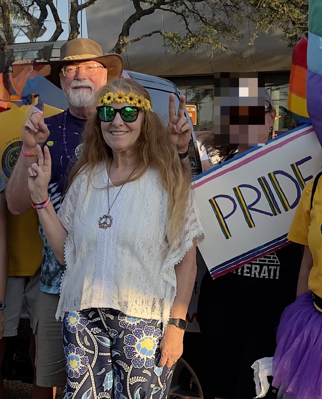 Terry in a sunflower headband and tie-dye, flashing peace signs beside her husband Clif in a wide-brimmed hat, at a Pride parade.