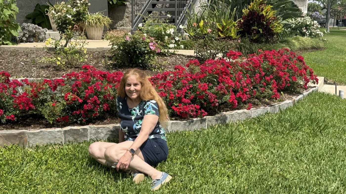Terry kneeling on the lawn in front of a long flowerbed of bright red Drift roses in full bloom.